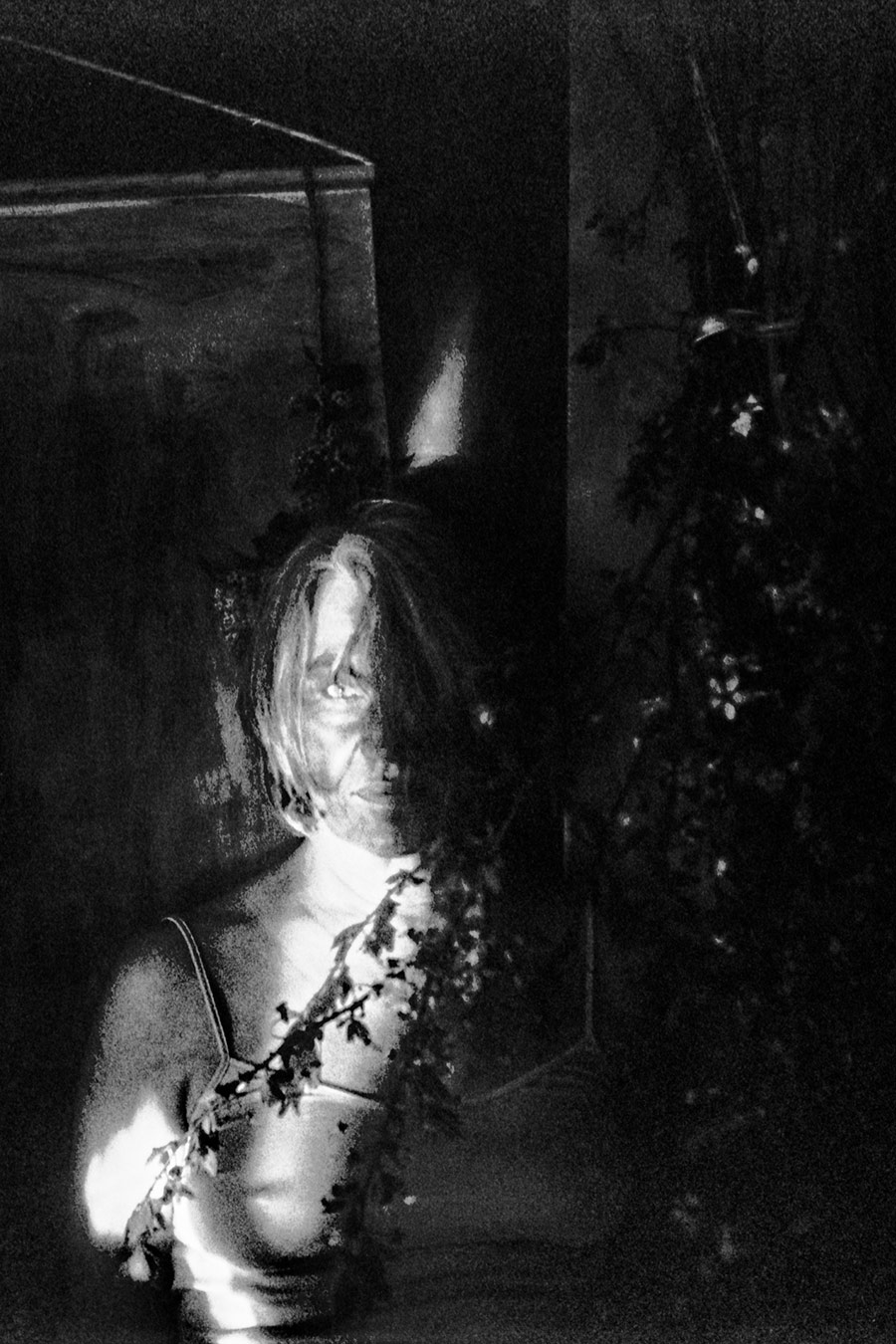 Nahaufnahme einer Frau im starken Kontrastlicht, Gesicht teilweise von Haaren und Zweigen verdeckt, in körnigem Schwarzweiß.