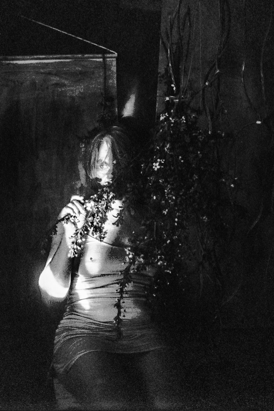 Frau im Sitzen in einem dunklen Raum, mit Zweigen in der Hand, partiell stark beleuchtet, umgeben von Gefäßen und Schatten.