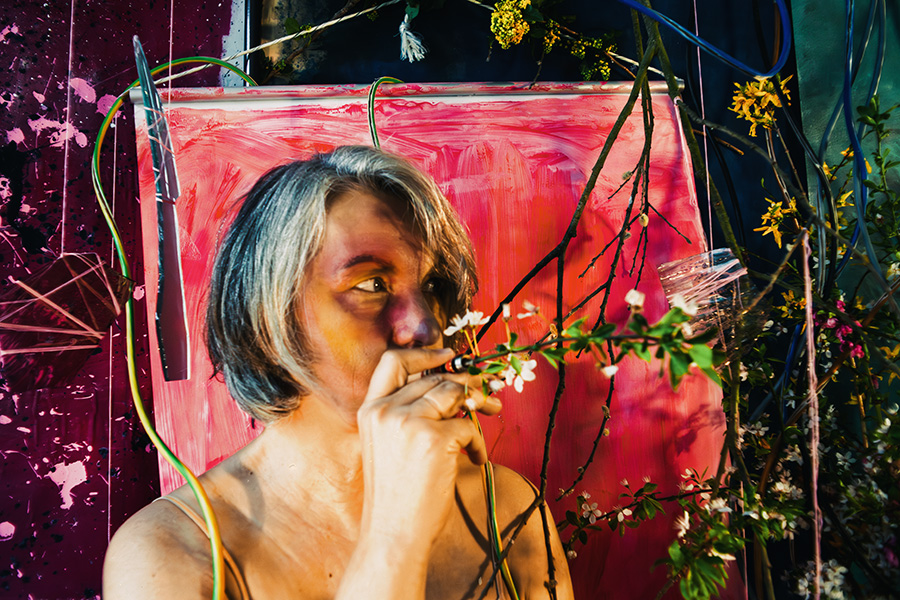 Fotokunst Greisenmädchen, Frau riecht an einem Blütenzweig mit seitlichem Blick, pinkes und rotes Gemälde dahinter, Fotografin Leipzig