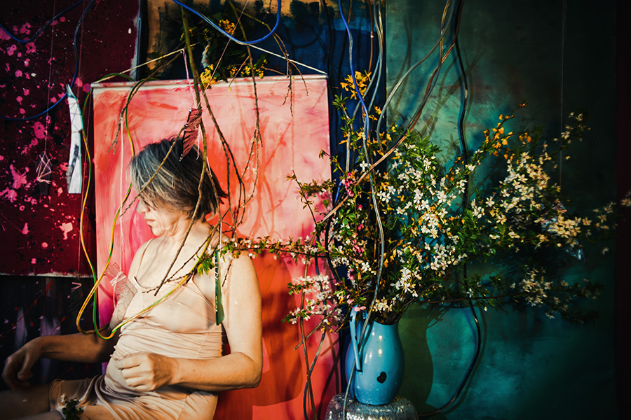 Fotokunst Greisenmädchen, Frau sitzt seitlich und schaut nach unten, große Blüten- und Kabelinstallation dahinter, Fotografin Leipzig