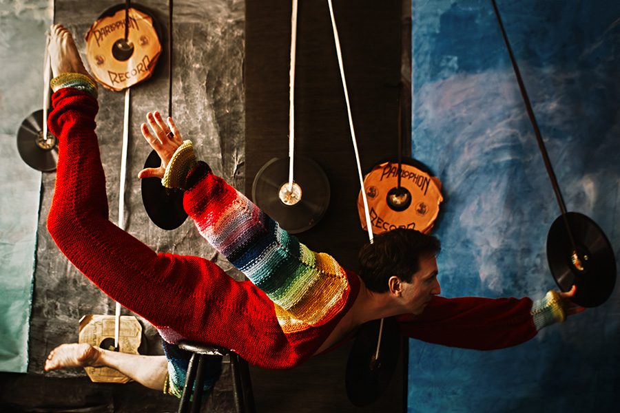 Farbfoto von oben: Mann in rotem Regenbogen-Strickanzug liegt horizontal ausgestreckt, Arme weit geöffnet, Schallplatten und Schnüre ringsum, Vogelperspektive.