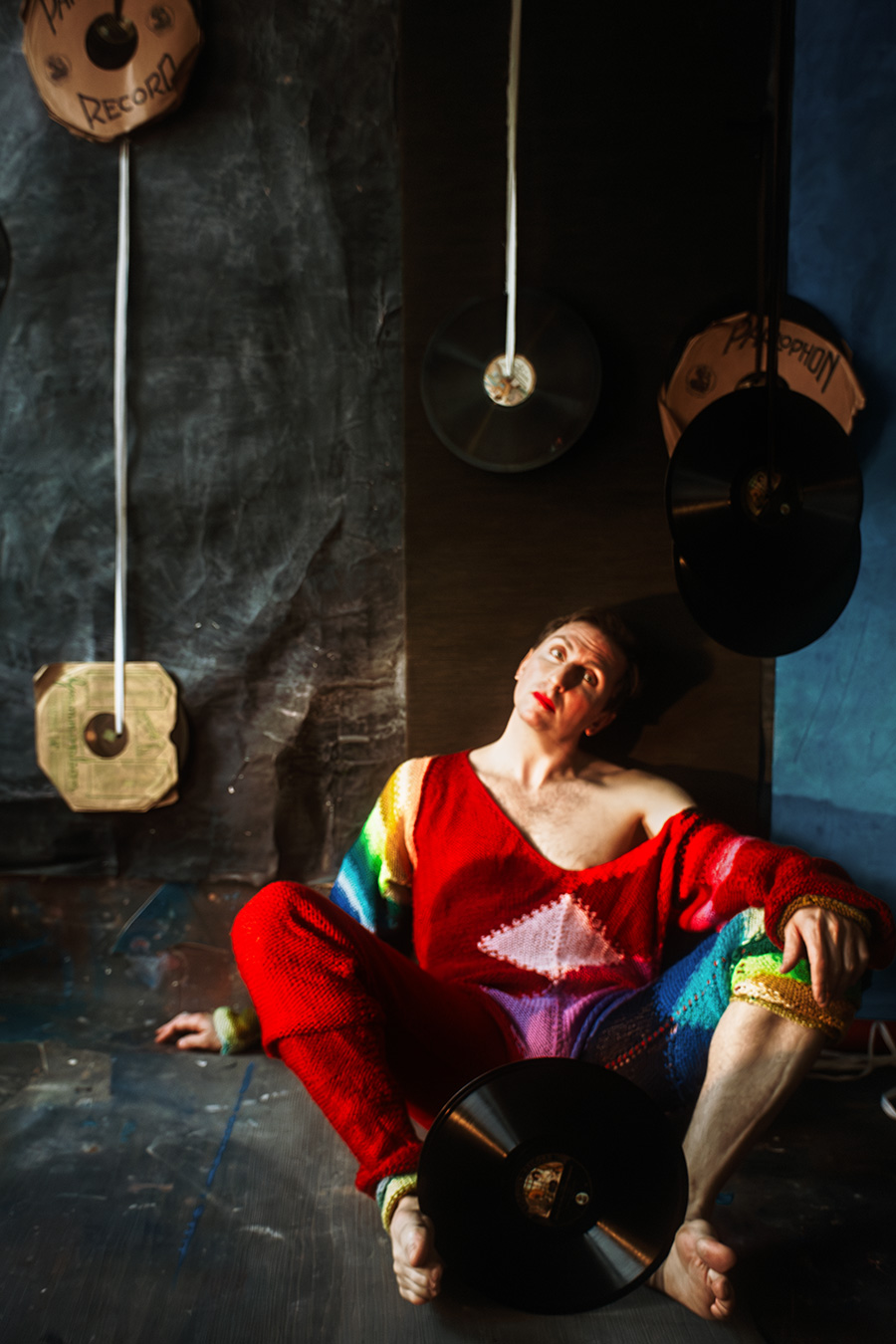 Mann in rotem Regenbogen-Strickanzug sitzt auf dem Boden, Blick nach oben, umgeben von hängenden Schallplatten, warmes Seitenlicht.