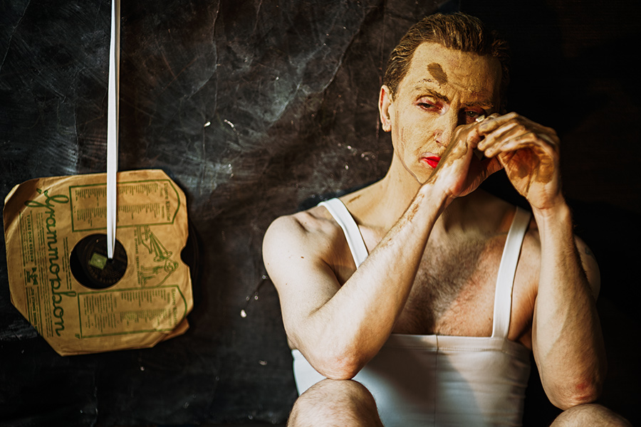 Farbfoto: Mann in weißem Trikot, Gesicht mit Ton bedeckt, reibt sich mit beiden Händen Farbe ins Gesicht, Blick gesenkt, Schallplatte im Hintergrund.