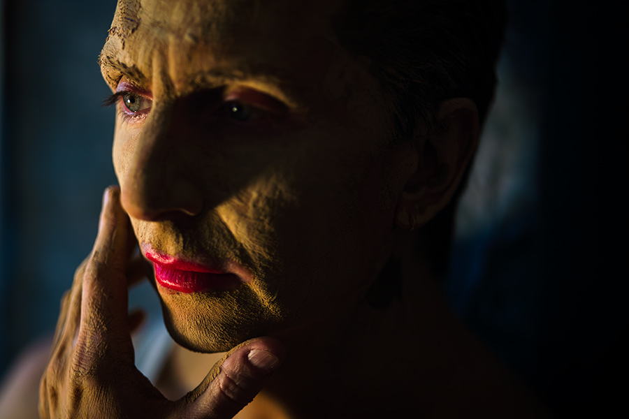 Nahaufnahme, Mann mit tonbedecktem Gesicht, Hände halten Kopf und Hals, Augen geschlossen, Kopf zurück, roter Mund, warmes Licht.