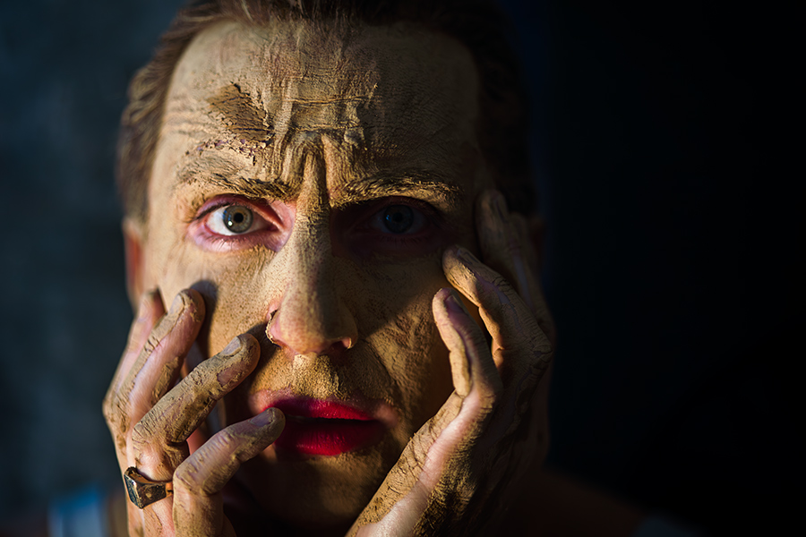 Farbfoto mit Bewegungsunschärfe: Nahaufnahme, Gesicht mit Ton bedeckt, Hände in Bewegung vor dem Gesicht, ein Auge geöffnet, warmes Licht, dunkler Hintergrund.