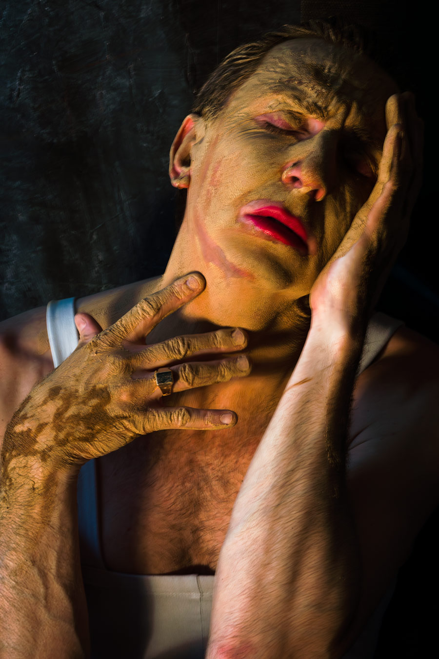 Farbfoto: Nahaufnahme, Gesicht mit Ton bedeckt, beide Hände halten das Gesicht, beide Augen offen, direkter Blick, roter Mund, dunkler Hintergrund.