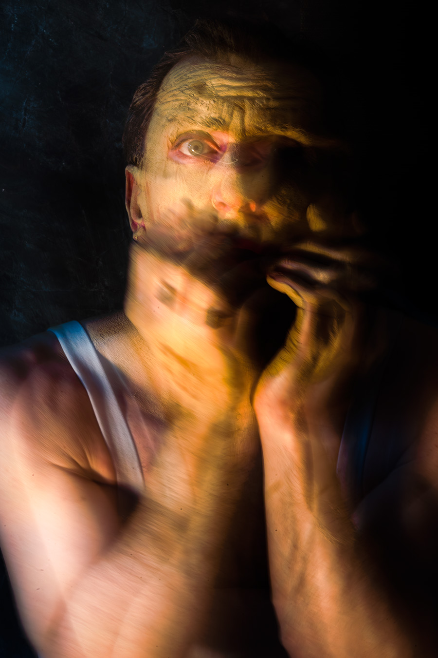 Farbfoto: Nahaufnahme, Mann mit tonbedecktem Gesicht und rotem Lippenstift, eine Hand an der Wange, ein Auge halb geöffnet, dunkler Hintergrund.