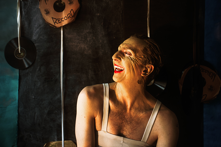 Farbfoto: Mann in weißem Trikot, Gesicht mit Erde gealtert, lacht weit auf, Kopf zurückgeworfen, rote Lippen, Schallplatten im Hintergrund.