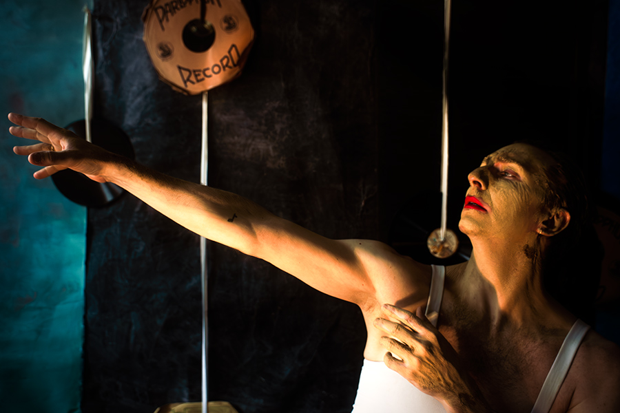 Farbfoto: Mann in weißem Trikot, Gesicht mit Ton bedeckt, streckt einen Arm weit aus, Kopf zurückgeworfen, rote Lippen, Schallplatten im Hintergrund.