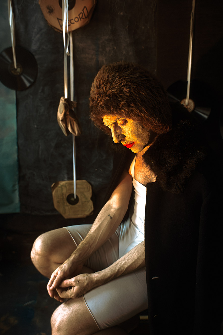 Farbfoto: Mann in weißem Trikot, Gesicht mit Erde bedeckt und gealtert, trägt dunkle Pelzkappe, sitzt erschöpft, Blick gesenkt, Schallplatten hängen im Hintergrund.