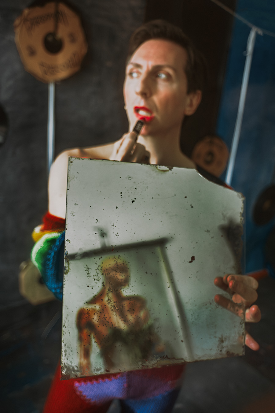 Mann in rotem Strickanzug schminkt sich mit rotem Lippenstift, hält einen alten beschädigten Spiegel, Reflexion darin kaum erkennbar.