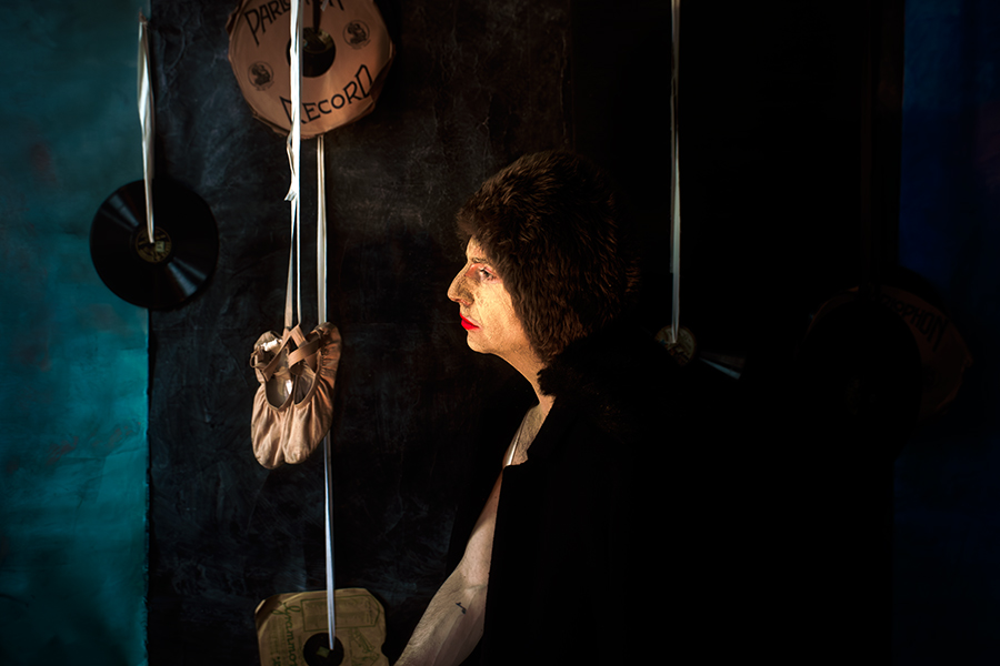 Mann in schwarzem Mantel, Gesicht mit Erde gealtert, steht im Profil, hält Spitzenschuh an einer Schnur, Blick nach vorn, Schallplatten im Hintergrund.