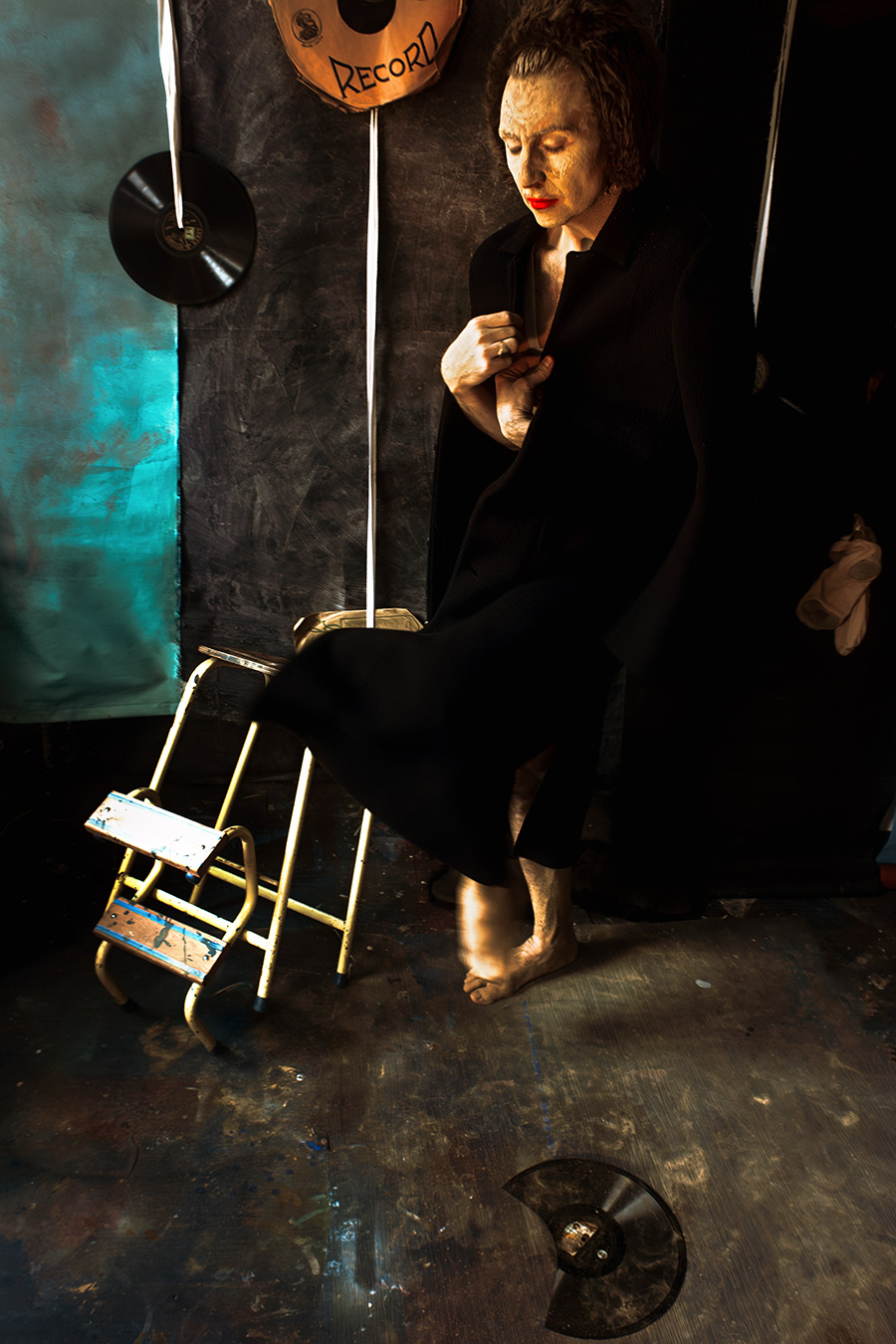 Mann in schwarzem Mantel, Gesicht durch Erdmaske gealtert, stützt ein Bein auf einen Hocker, Blick nach unten, rote Lippen, Schallplatten im Hintergrund.