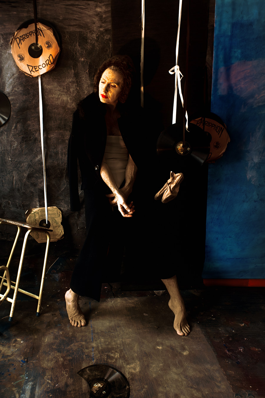 Farbfoto: Mann in schwarzem Mantel, Gesicht durch Erdmaske gealtert, steht barfuß, Hände gefaltet, Blick gesenkt, Spitzenschuhe hängen an Schnur, Schallplatten ringsum.