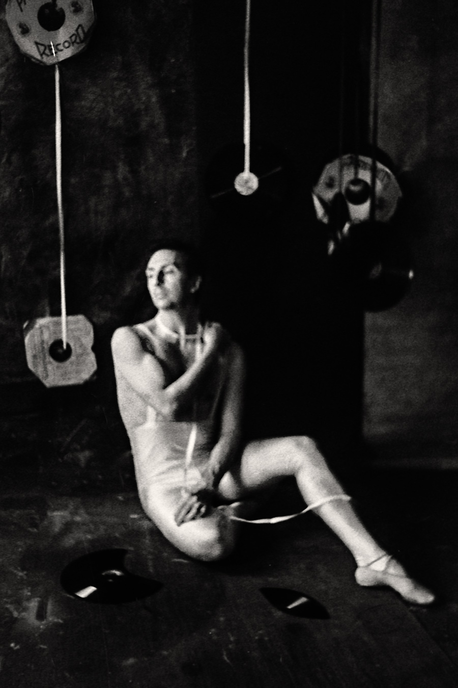 Fomapan 400 gepusht · Zenit, Stark gekörntes Schwarzweißfoto: Tänzer in weißem Trikot sitzt auf dem Boden, Blick leicht nach oben, Spitzenschuhe, Schallplatten im Hintergrund, weiches Licht.