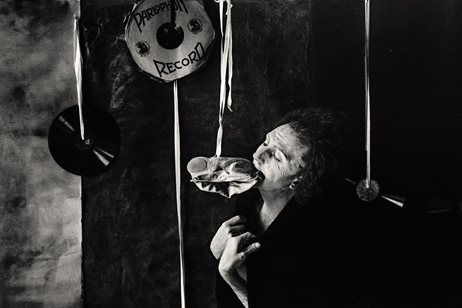Tmax 100 auf 800 gepusht, analoge Fotografie Leipzig, Schwarzweißfoto: Mann in dunklem Oberteil hält einen Spitzenschuh im Mund, Blick nach oben, gealterte Haut, Schallplatten im Hintergrund.