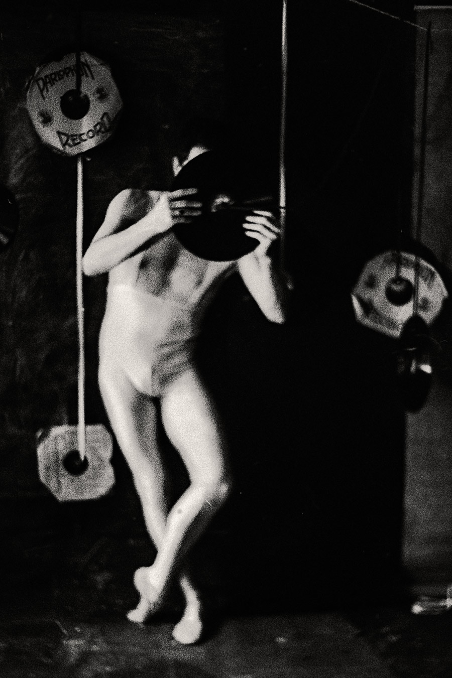 Fomapan 400 gepusht · Zenit, Stark gekörntes Schwarzweißfoto: Nackter Körper streckt Arm aus, Bein erhoben, zwischen hängenden Schallplatten, körnig und dunkel, fast abstrakt.
