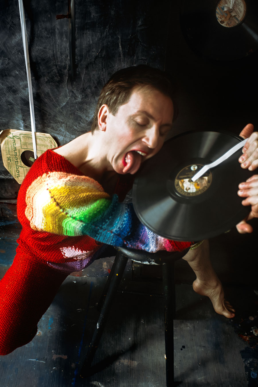 Farbfoto: Mann in rotem Regenbogen-Strickanzug reißt den Mund auf, Zunge herausgestreckt, Schallplatte nah am Gesicht, Körper in extremer Schräglage.