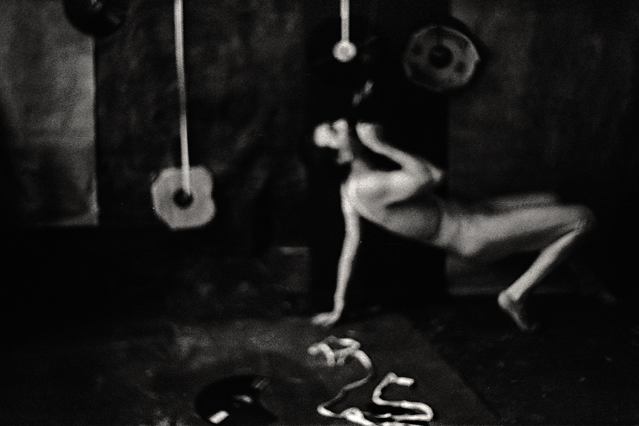 Fomapan 400 gepusht · Zenit, analoge fotografie,Stark gekörntes Schwarzweißfoto: Verschwommene stehende Figur in weißem Trikot, Bewegungsunschärfe, Spitzenschuhe auf dem Boden, körniges Dunkel.