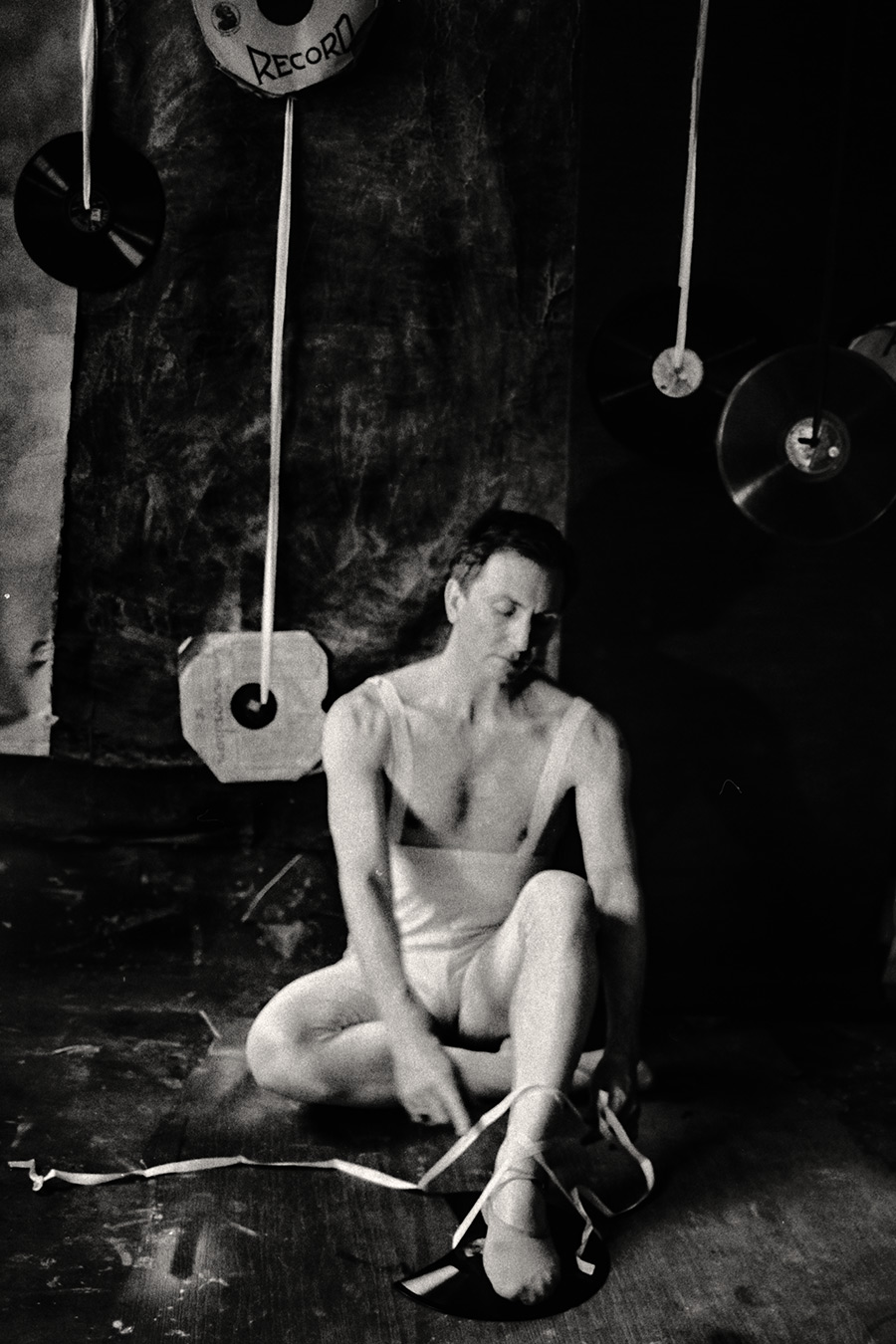 Fomapan 400 gepusht · Zenit, analoge fotografie,Schwarzweißfoto: Mann in weißem Trikot sitzt auf dem Boden, bindet seinen Spitzenschuh, Blick gesenkt, Schallplatten hängen im Hintergrund.
