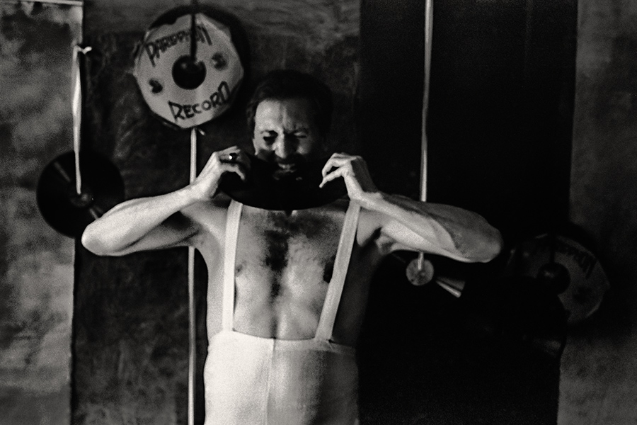 Fomapan 400 gepusht · Zenit, analoge fotografie, Schwarzweißfoto: Mann in weißem Trikot steht, zieht mit angespanntem Gesicht an den Trägern seines Trikots, Schallplatten im Hintergrund.