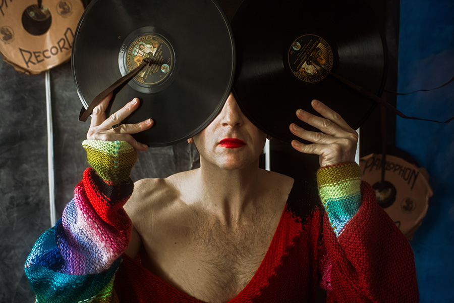 Mann in rotem Strickanzug hält zwei Schallplatten vor die Augen, rote Lippen sichtbar, Blick verborgen hinter schwarzem Vinyl.