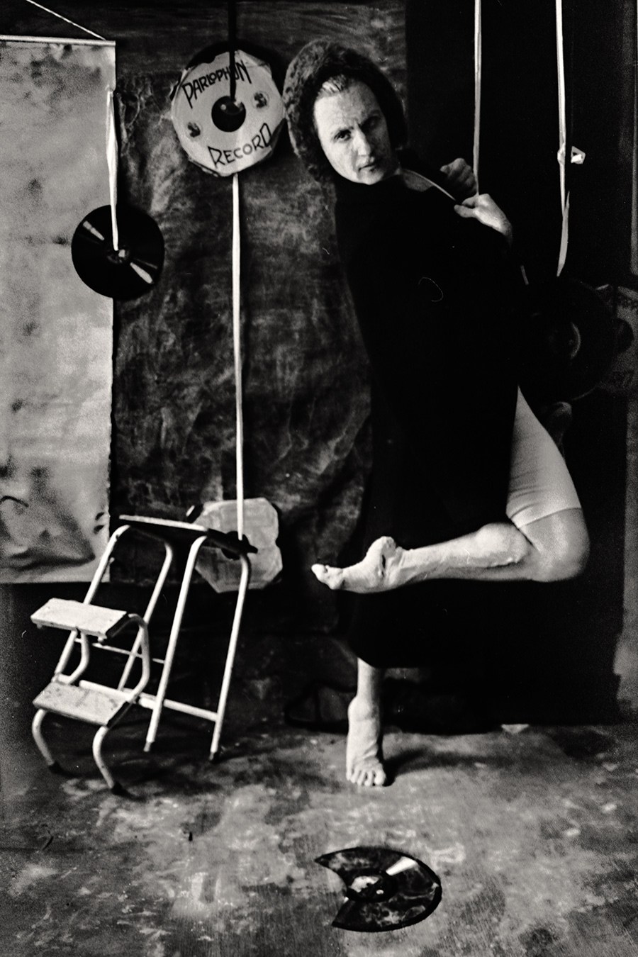 Tmax 100 auf 800 gepusht, analoge Fotografie Leipzig,balett, Mann in schwarzem Mantel hebt ein Bein hoch, barfuß, ein Hocker steht leer daneben, Schallplatten hängen im Hintergrund.