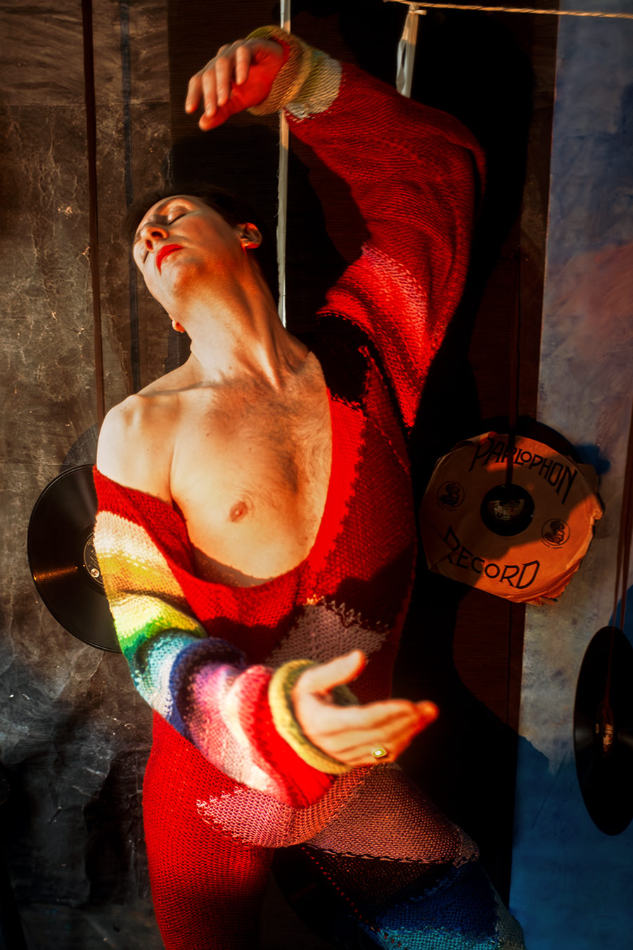 Mann in rotem Regenbogen-Strickanzug, Oberkörper frei, Kopf zurückgeworfen, Arm erhoben, Schallplatten und Schnüre im Hintergrund.