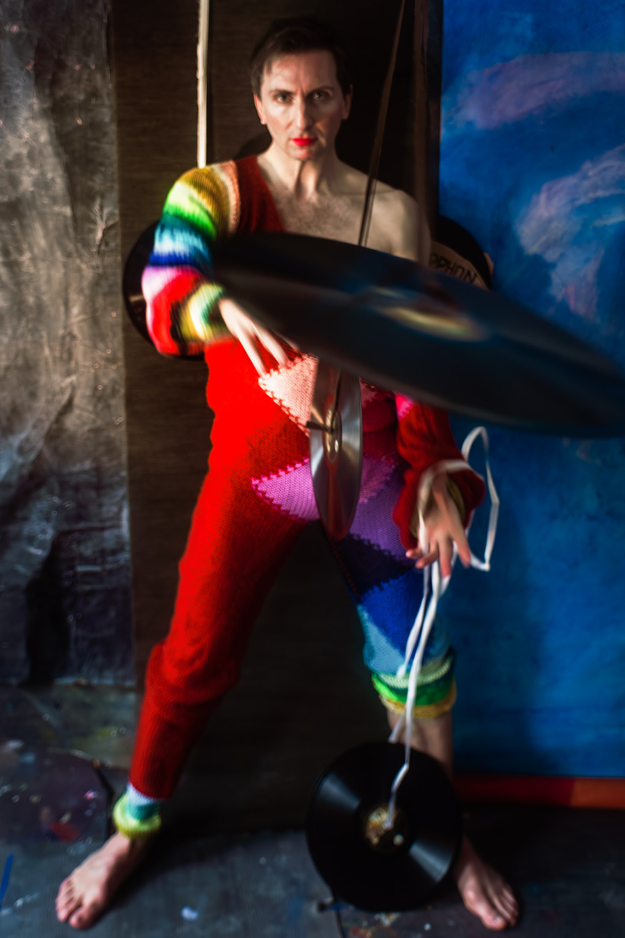 Farbfoto: Mann in rotem Regenbogen-Strickanzug steht aufrecht, hält Schallplatte im Arm, direkter Blick in die Kamera, rote Lippen, Seil in der Hand.