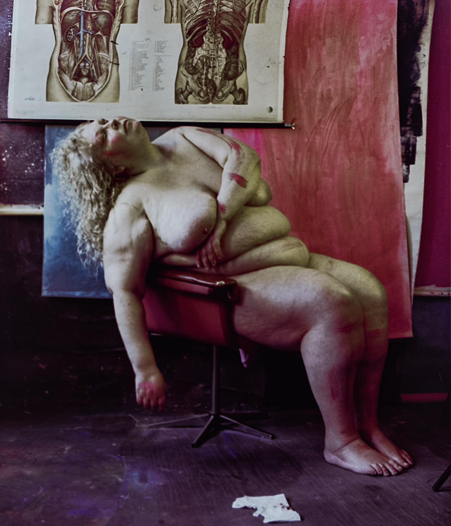 Nackte Frau liegt halb vom Stuhl gleitend, Kopf zurückgeworfen, lockiges Haar, wenige rote Farbspuren auf Körper, roter Vorhang und anatomische Tafeln im Hintergrund. Aktfotografie, Nude. Dicker Körper einer Frau