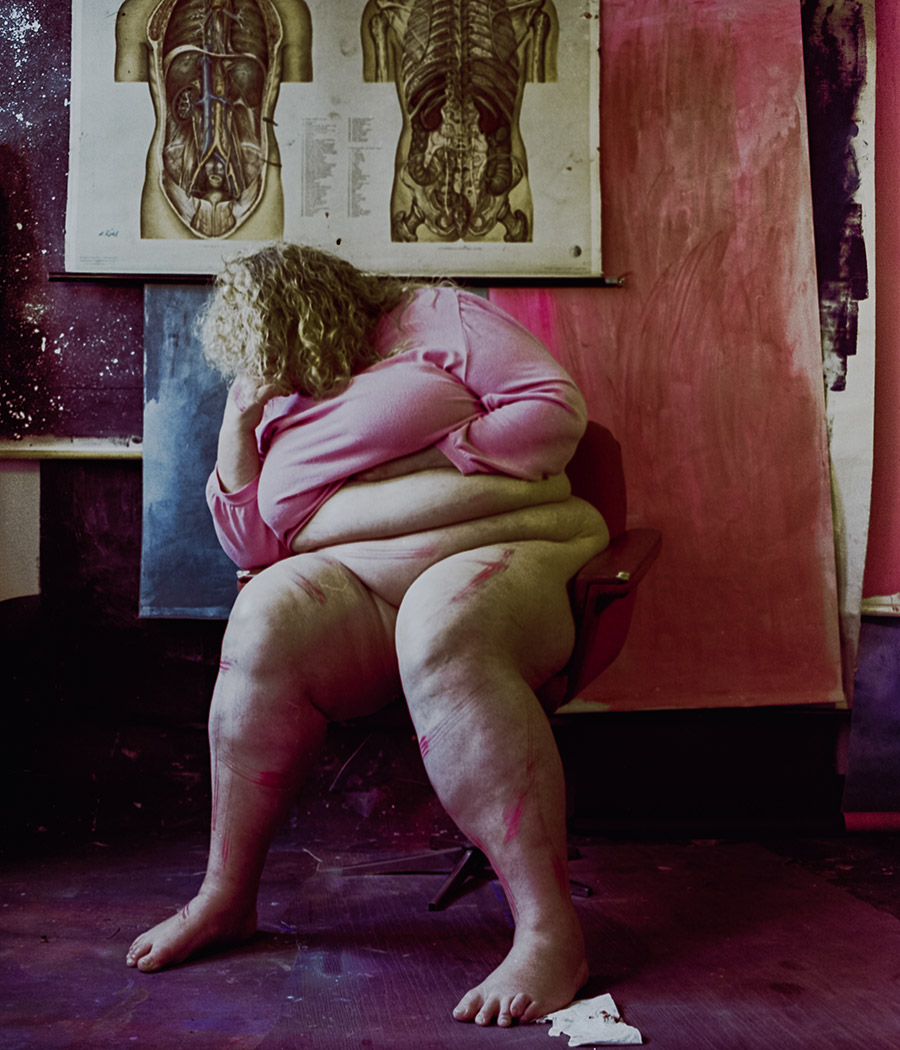 Analoges Farbfoto: Frau sitzt zusammengekauert auf Stuhl, pinkes Shirt, Gesicht in Armen verborgen, rote Farbstriche auf Beinen, roter Hintergrund, anatomische Tafel oben. Aktfotografie, Nude