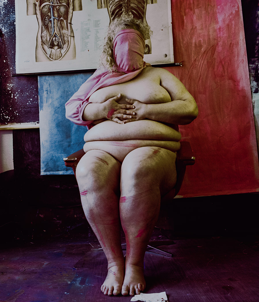 Analoges Farbfoto: Nackte Frau liegt halb vom Stuhl gleitend, Kopf zurückgeworfen, lockiges Haar, wenige rote Farbspuren auf Körper, roter Vorhang und anatomische Tafeln im Hintergrund. Aktfotografie, Nude. Dicker Körper einer Frau