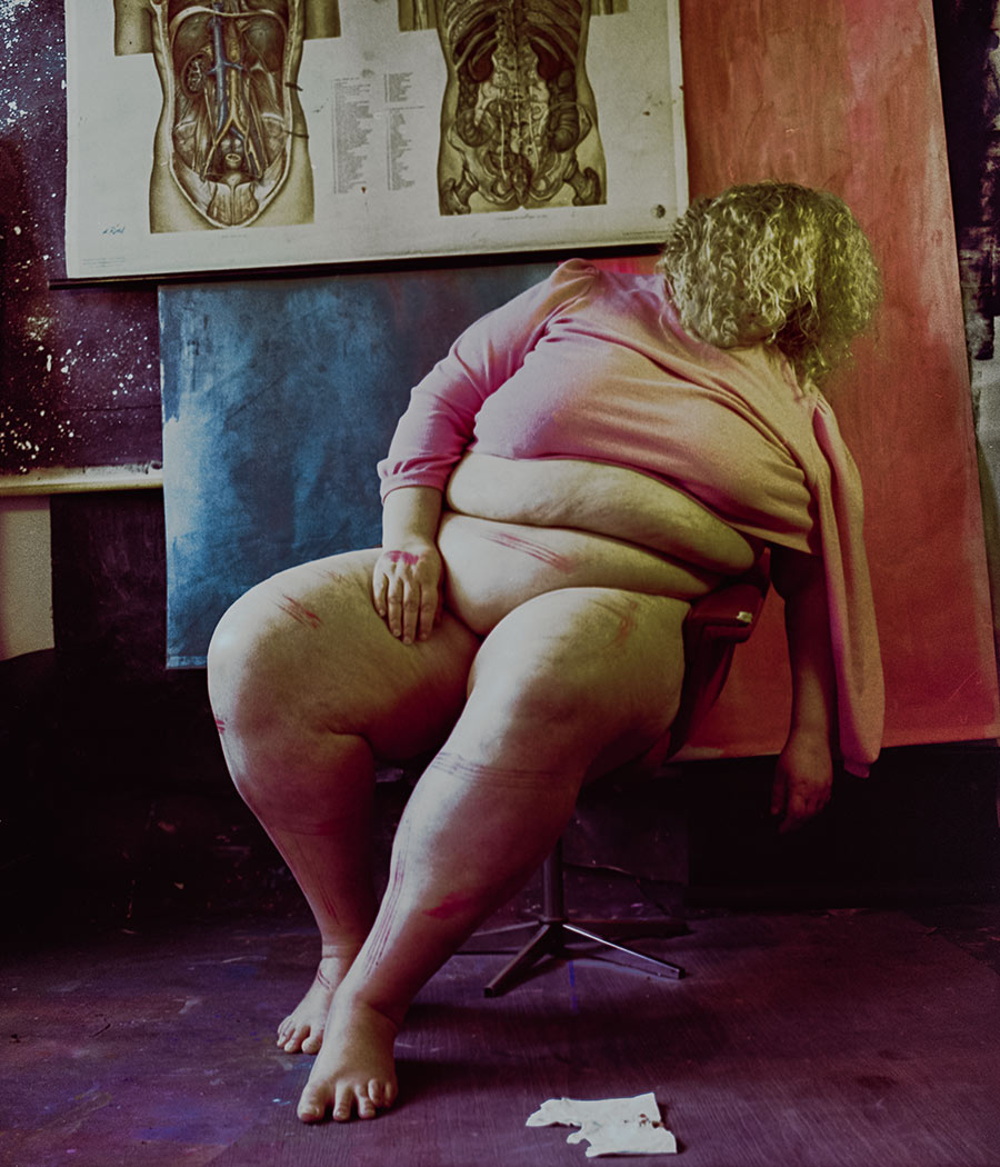 Analoges Farbfoto: Frau von hinten auf Stuhl sitzend, pinkes Shirt, Gesicht abgewandt, rote Farbstriche auf Beinen und Hüfte, anatomische Tafel im Hintergrund. Aktfotografie, Nude