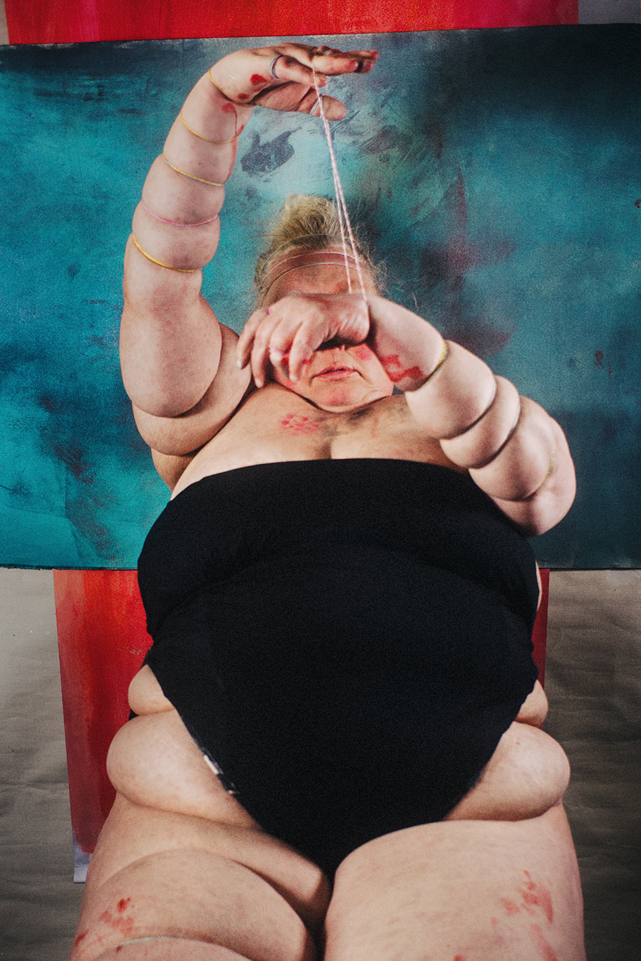 Sitzende Figur, eingeschnürter Arm steil nach oben gereckt, Faden an den Fingern, Gesicht halb hinter der anderen Hand verborgen – inszeniertes Selbstporträt Antje Kröger Berlin, Serie Mehrenge