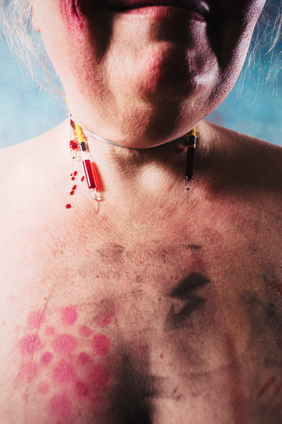 Nahaufnahme Hals und Brust, zwei Spritzen als Halsschmuck an dünnem Faden, Bluttropfen auf der Haut, pinke Druckpunkte auf der Brust – konzeptuelle Selbstporträt-Fotografie Antje Kröger Leipzig, Serie Mehrenge