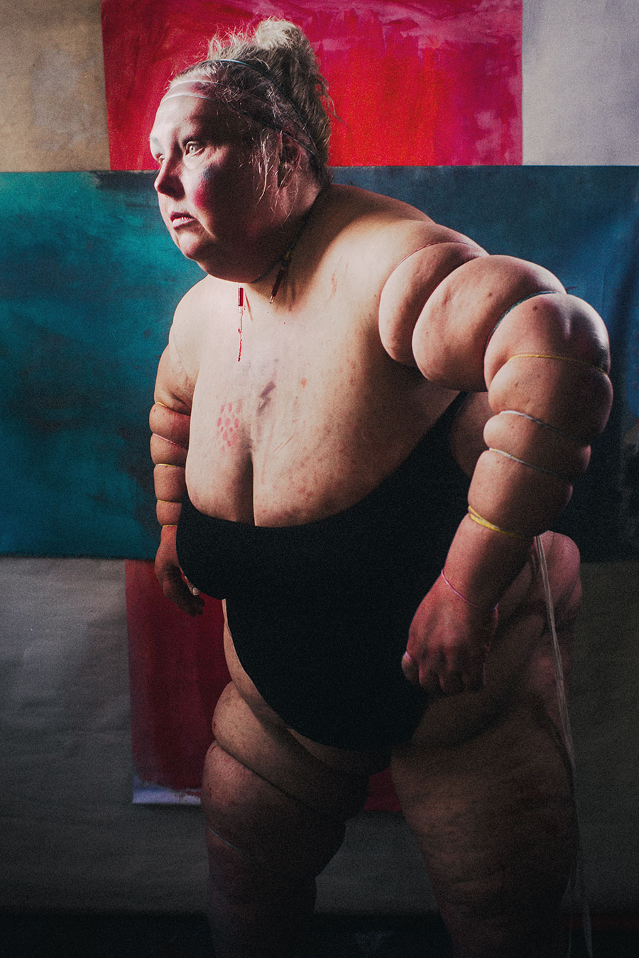 Ganzkörper-Selbstporträt im schwarzen Einteiler, ein Arm seitlich ausgestreckt und von Gummibändern eingeschnürt, rote Linie auf dem Rücken, dreiteiliger gemalter Hintergrund – Fotokunst Antje Kröger Leipzig, Serie Mehrenge