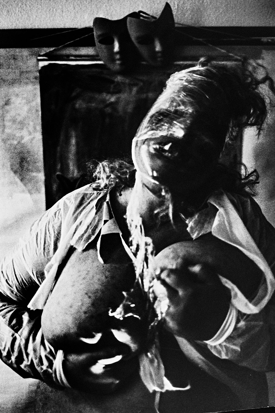 analoge fotokunst leipzig, Analogfoto auf Fomapan 400 Ortho, um eine Stufe gepusht, stark körnig. Eine dicke Frau mit langen Haaren drückt einen dunklen Maskenkopf an ihren Körper. Ihr eigenes Gesicht ist durch weißen Kunststoffstoff verzerrt und kaum erkennbar. Im Hintergrund ein Metallgestell mit zwei schwarzen Masken.