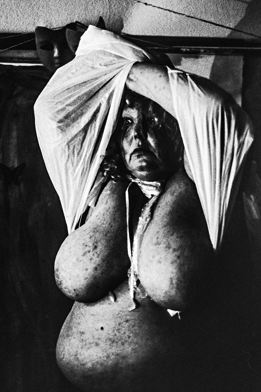 analoge fotokunst leipzig, Analogfoto auf Fomapan 400 Ortho, um eine Stufe gepusht, stark körnig, leicht unscharf. Eine dicke Frau mit langen dichten lockigen Haaren im Seitenprofil, die Arme vor dem Körper verschränkt. Im Hintergrund ein dunkles Metallgestell mit einer schwarzen Maske.