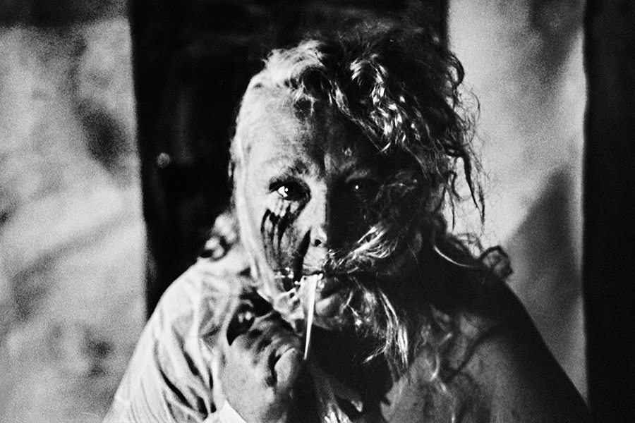 analoge fotokunst leipzig, Analogfoto auf Fomapan 400 Ortho, um eine Stufe gepusht, stark körnig. Nahaufnahme des Gesichts einer dicken Frau mit lockigem Haar, das vollständig schwarz bemalt ist. Die Augen sind hell und weit geöffnet, der Mund leicht geöffnet, ein weißer dünner Faden hängt aus dem Mund.