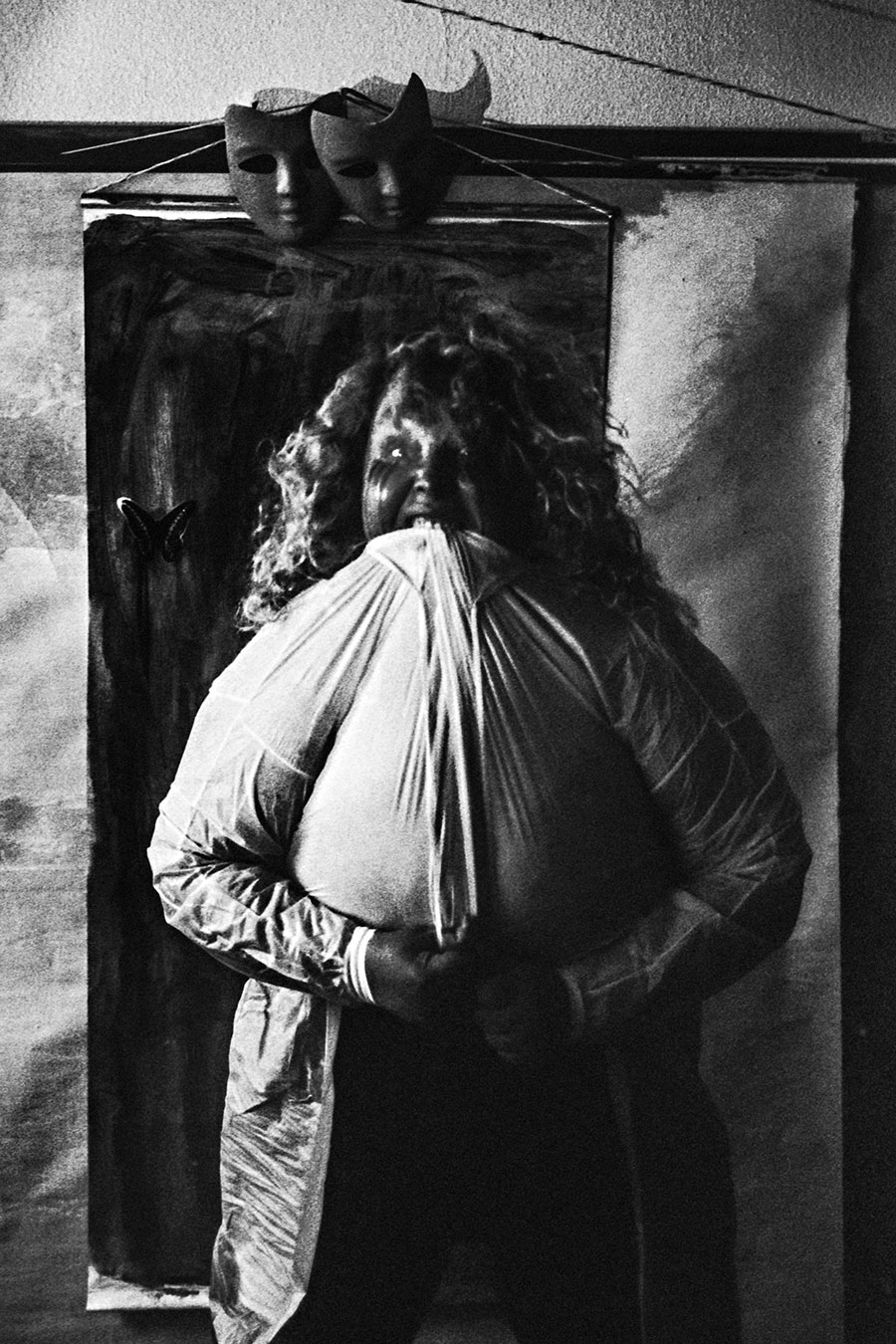 analoge fotokunst leipzig, Analogfoto auf Fomapan 400 Ortho, um eine Stufe gepusht, stark körnig. Eine dicke Frau mit langen Haaren drückt einen dunklen Maskenkopf an ihren Körper. Ihr eigenes Gesicht ist durch weißen Kunststoffstoff verzerrt und kaum erkennbar. Im Hintergrund ein Metallgestell mit zwei schwarzen Masken.