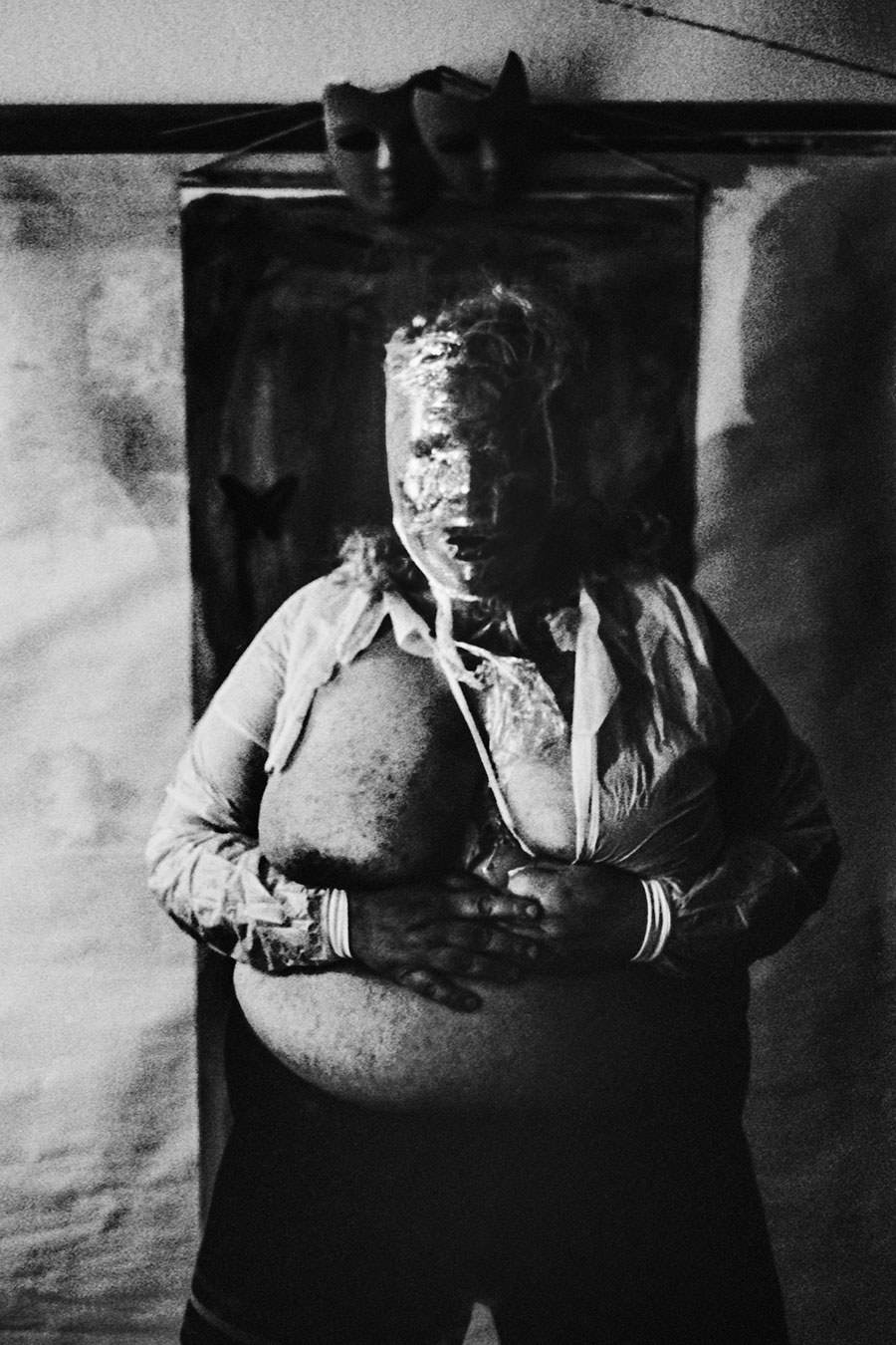 analoge fotokunst leipzig, Analogfoto auf Fomapan 400 Ortho, um eine Stufe gepusht, stark körnig. Eine dicke Frau steht frontal, beide Hände auf dem runden Bauch. Ihr Gesicht ist mit einer glänzenden hellen Substanz versiegelt. Im Hintergrund ein Metallgestell mit einer schwarzen Maske, auf der dunkel bemalten Fläche ein kleiner schwarzer Schmetterling