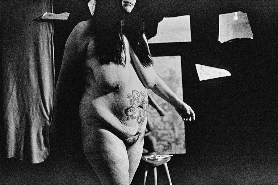 Analoge Aktfotografie Schwarz-Weiß, dicke Frau nackt stehend, großes Tattoo auf dem Bauch sichtbar, Kopf angeschnitten, dunkler Hintergrund mit zerrissenen Papierfetzen und Leinwand, Holzboden, körnige Filmfotografie, Ganzkörperaufnahme, Fotokunst.