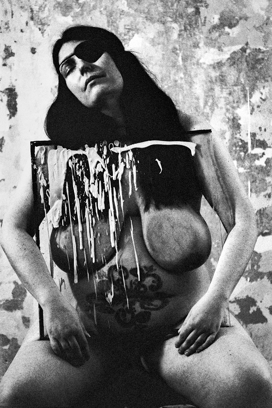 Analoge Aktfotografie Schwarz-Weiß, dicke Frau mit schwarzer Augenklappe sitzt nackt auf einem Stuhl, vor ihrem Körper ein mit weißer Farbe bespritztes schwarzes Gemälde, verwitterte Wand im Hintergrund, körnige Filmfotografie, Fotokunst.