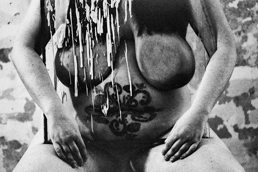 Analoge Aktfotografie Schwarz-Weiß, Detailaufnahme des nackten Oberkörpers einer dicken Frau, großes dekoratives Tattoo auf dem Bauch, über den Körper läuft weiße Farbe in langen Tropfen herab, dahinter ein dunkles bespritztes Gemälde, verwitterte Wand im Hintergrund, körnige Filmfotografie, Fotokunst.