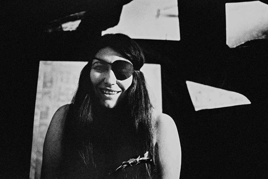 Analoge Schwarz-Weiß-Fotografie, Frau mit schwarzer Augenklappe und langen dunklen Haaren lacht, bekleidet, dunkler Hintergrund mit zerrissenen Papierfetzen, körnige Filmfotografie, Behind the Scenes, Fotokunst.