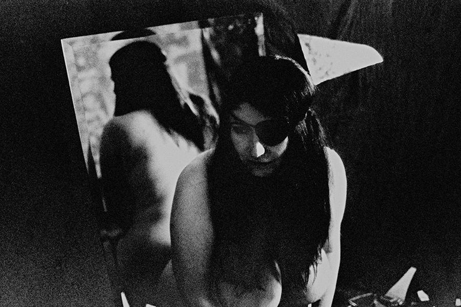 Analoge Aktfotografie Schwarz-Weiß, dicke Frau mit schwarzer Augenklappe und langen dunklen Haaren nackt, nach unten blickend, hinter ihr ein großer Abzug mit Silhouette desselben nackten Körpers, stark körnig, dramatische Lichtsetzung, Fotokunst.