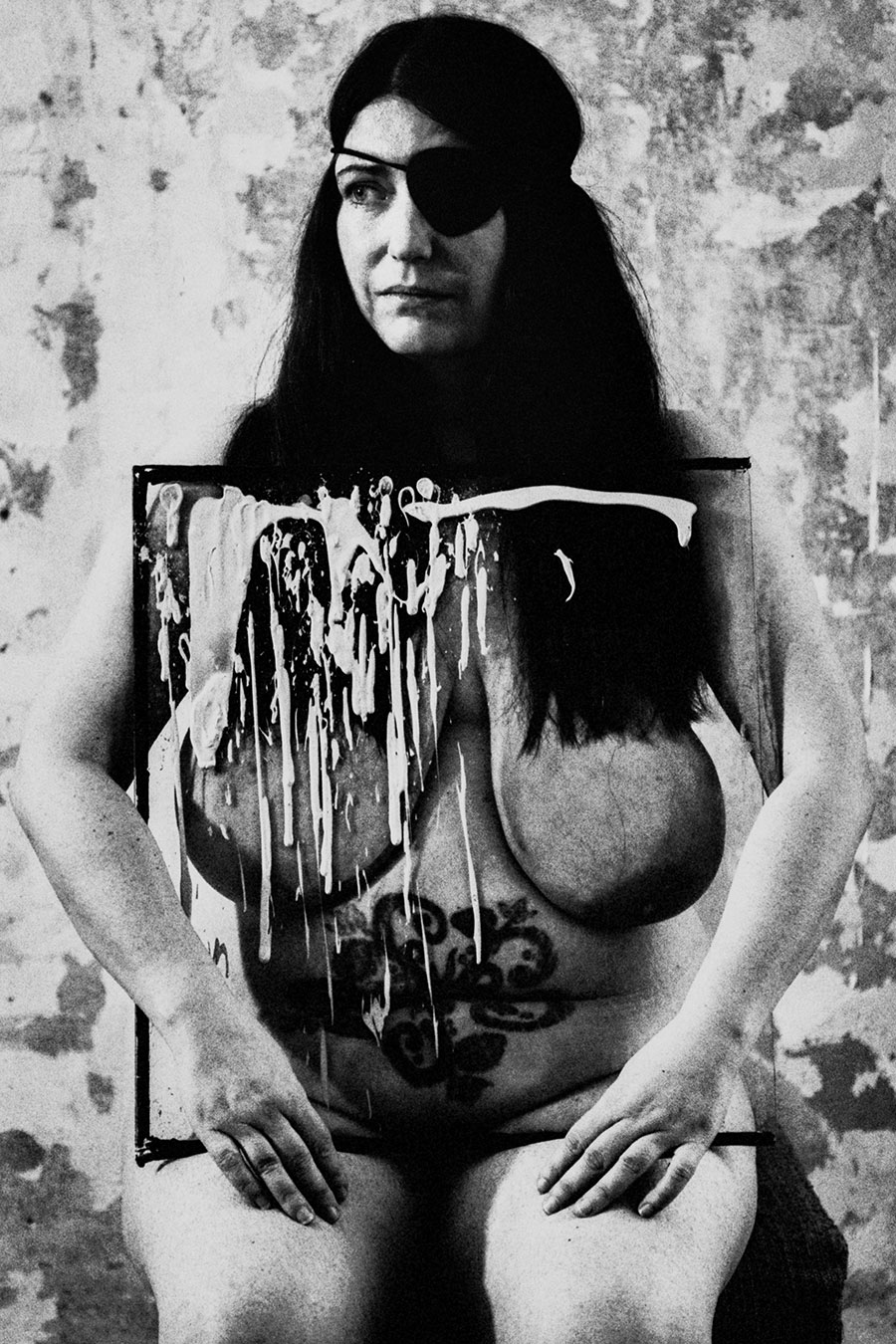 Analoge Aktfotografie Schwarz-Weiß, dicke Frau mit schwarzer Augenklappe und langen schwarzen Haaren sitzt frontal nackt auf Stuhl, Hände auf den Oberschenkeln, vor dem Körper ein mit weißer Farbe bespritztes Gemälde mit Tattoomotiven, verwitterte Wand, körnige Filmfotografie, Fotokunst.