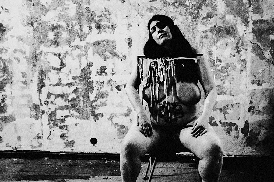 Analoge Aktfotografie Schwarz-Weiß, dicke Frau mit schwarzer Augenklappe sitzt nackt auf Stuhl, Kopf zurückgelehnt, vor dem Körper ein mit weißer Farbe bespritztes Gemälde, stark verwitterte Wand, Weitwinkelaufnahme, körnige Filmfotografie, Fotokunst.