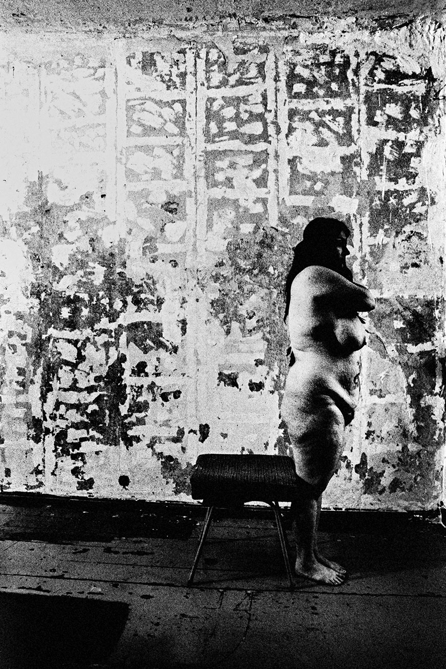 Analoge Aktfotografie Schwarz-Weiß, dicke Frau nackt im Seitenprofil stehend, Arme vor der Brust verschränkt, stark abgeblätterte Wand im Hintergrund, Holzboden, Hocker, Ganzkörperaufnahme, körnige Filmfotografie, Fotokunst.