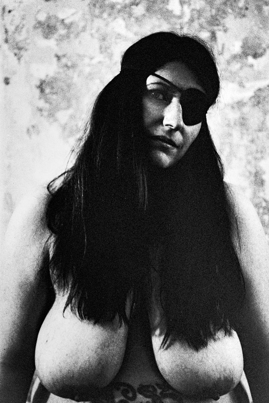 Analoge Aktfotografie Schwarz-Weiß, dicke Frau mit langen schwarzen Haaren und schwarzer Augenklappe, Nahaufnahme Porträt, nackter Oberkörper mit großen Brüsten, Tattoo angedeutet, verwitterte Wand, körnige Filmfotografie, Fotokunst.